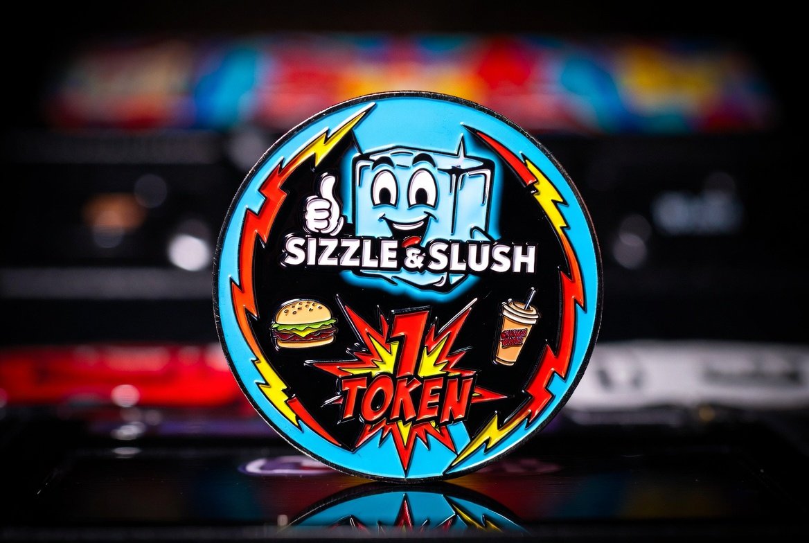 Sizzle & Slush 1 Token — the arcade coin standing on a cabinet: the smiling ice cube mascot over the name 'Sizzle & Slush,' a burger and a slush cup flanking a red and yellow '1 Token' burst ringed with lightning